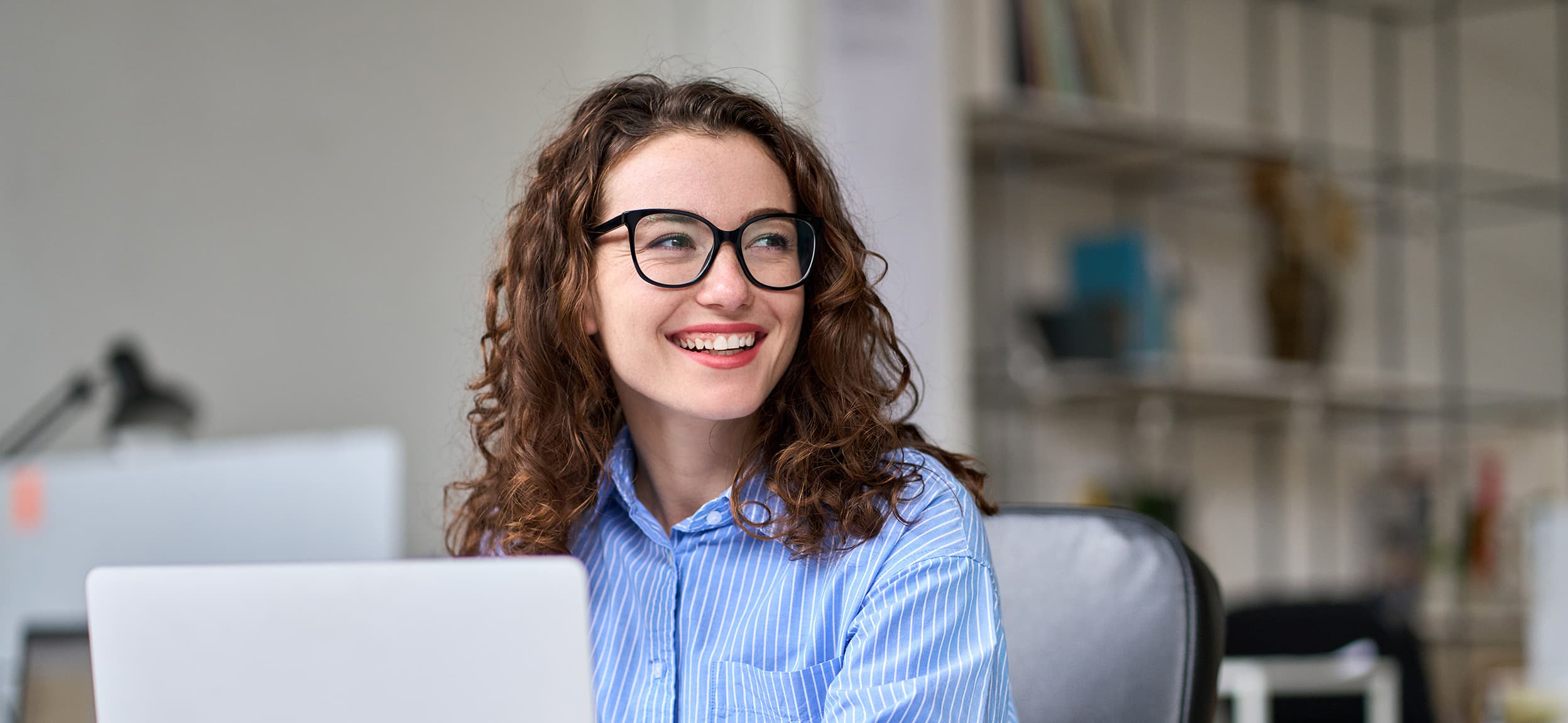 A professional smiles while working on computer and sitting in an office setting.